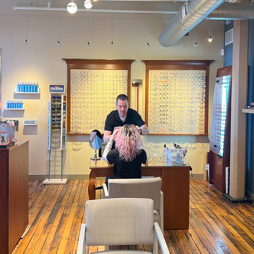 wide angle shot of an optical shop staff member working with a customer trying on glasses with optical shop displays behind them at the optical shop in Northampton MA